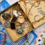 A vintage-style jewelry box filled with antique gold-toned necklaces, pendants, and charms, including a green stone pendant and coin-like charms, set against a blue patterned textile background.