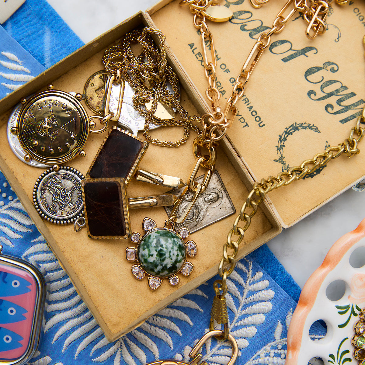 A vintage-style jewelry box filled with antique gold-toned necklaces, pendants, and charms, including a green stone pendant and coin-like charms, set against a blue patterned textile background.