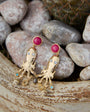 Gold earrings with pinkish red gemstones shaped like an octopus on a stone surface 