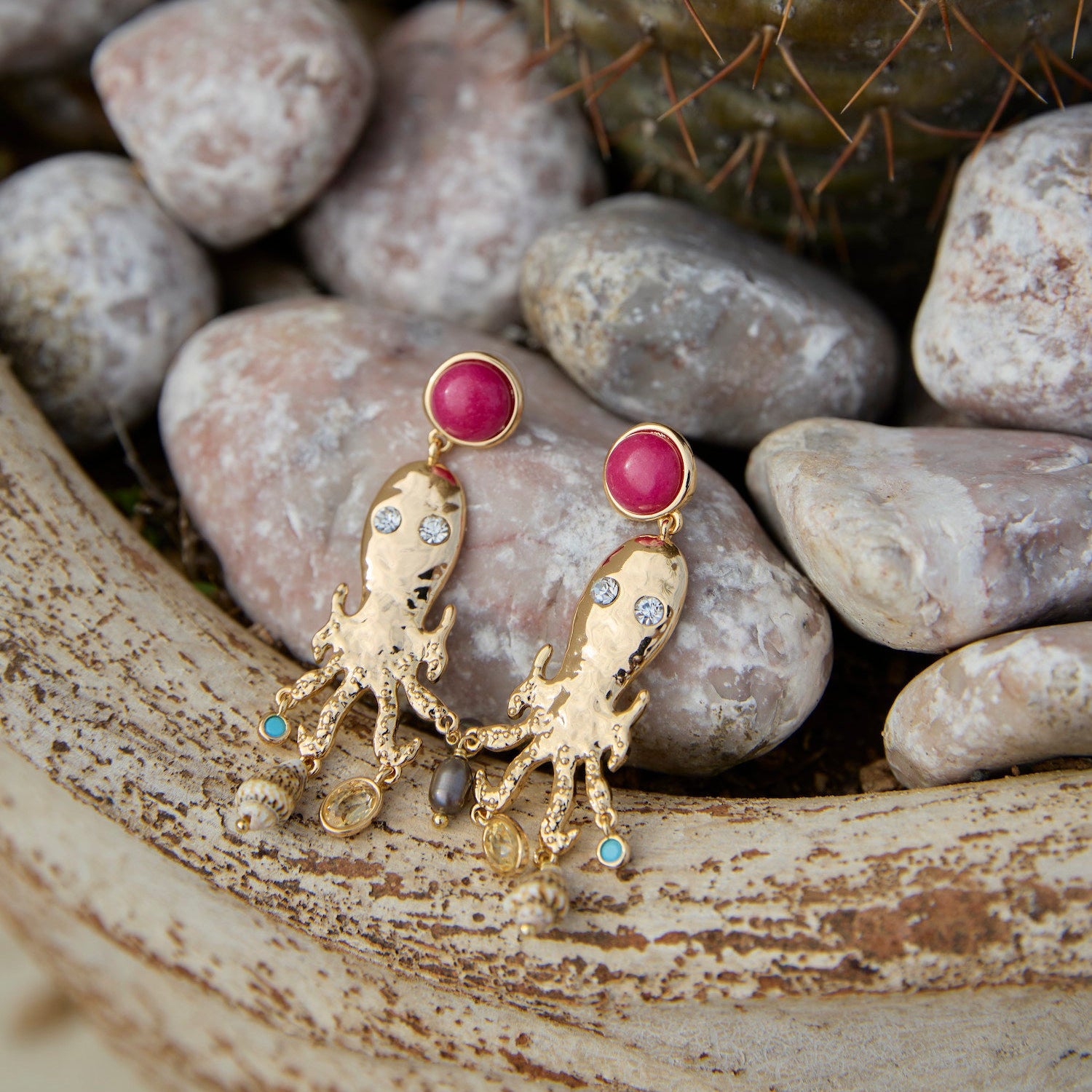 Gold earrings with pinkish red gemstones shaped like an octopus on a stone surface 
