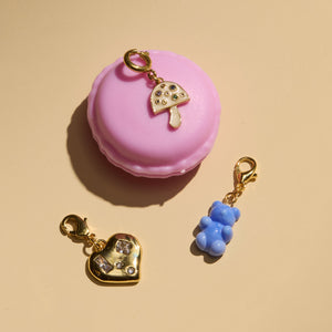 Crystal and Gold Heart Charm, Glass Blue Gummy Bear Charm, and Enamel and Crystal Mushroom Charm Staged on Pink Macaroon