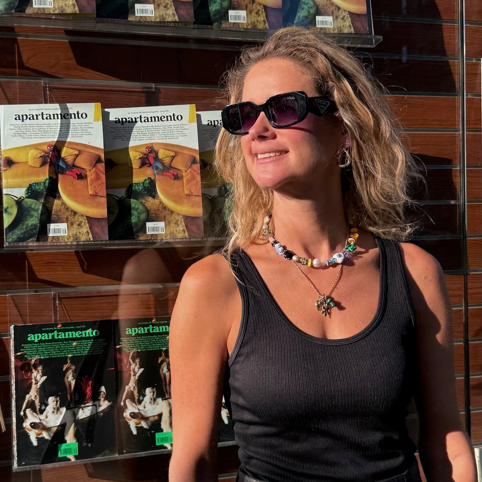 Woman standing in front of a display of 'Apauro Lamento' books or packaging wearing beaded strand necklace and charm necklace