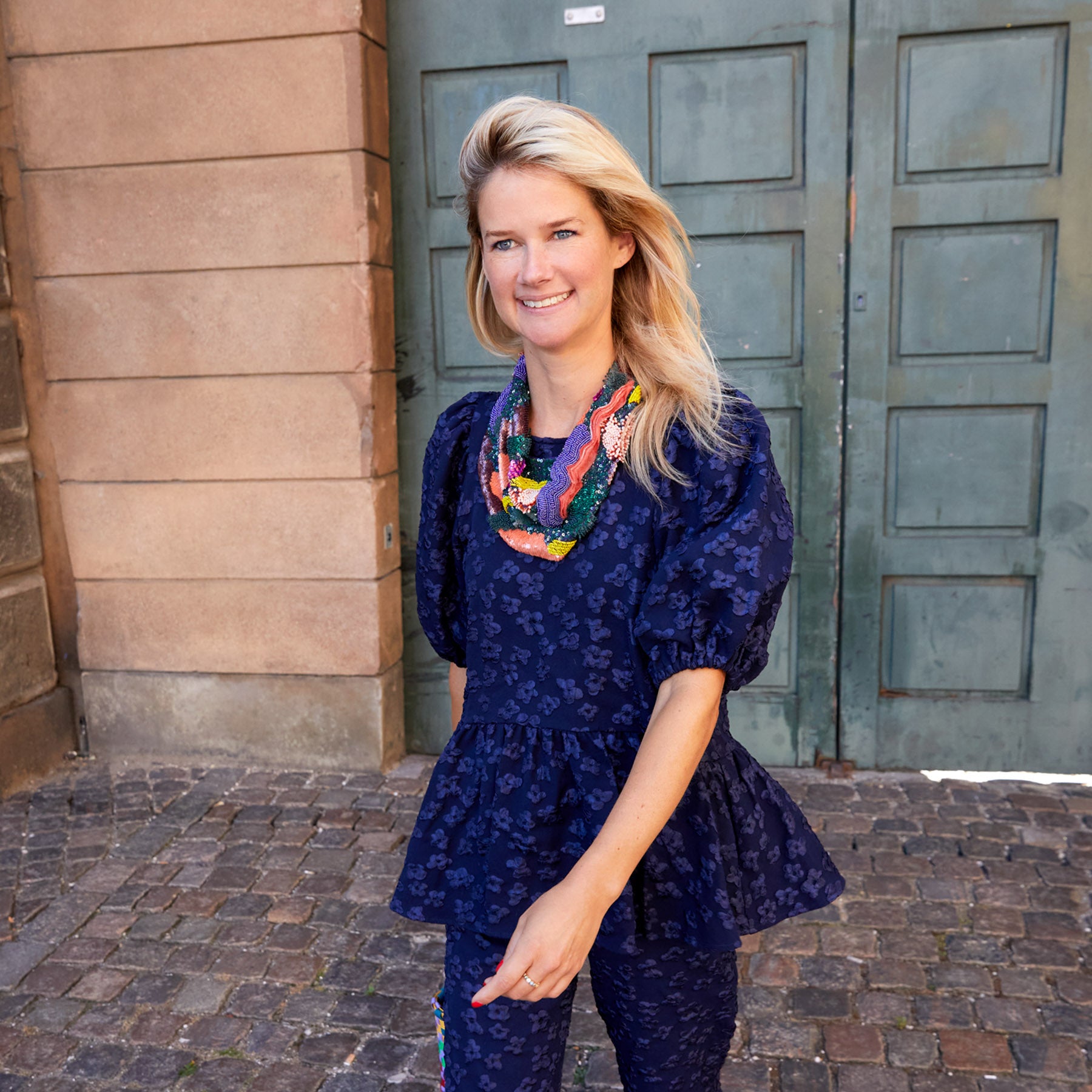 Maggie in a blue floral top and pants standing in front of a building wearing a multi-colored scarf necklace
