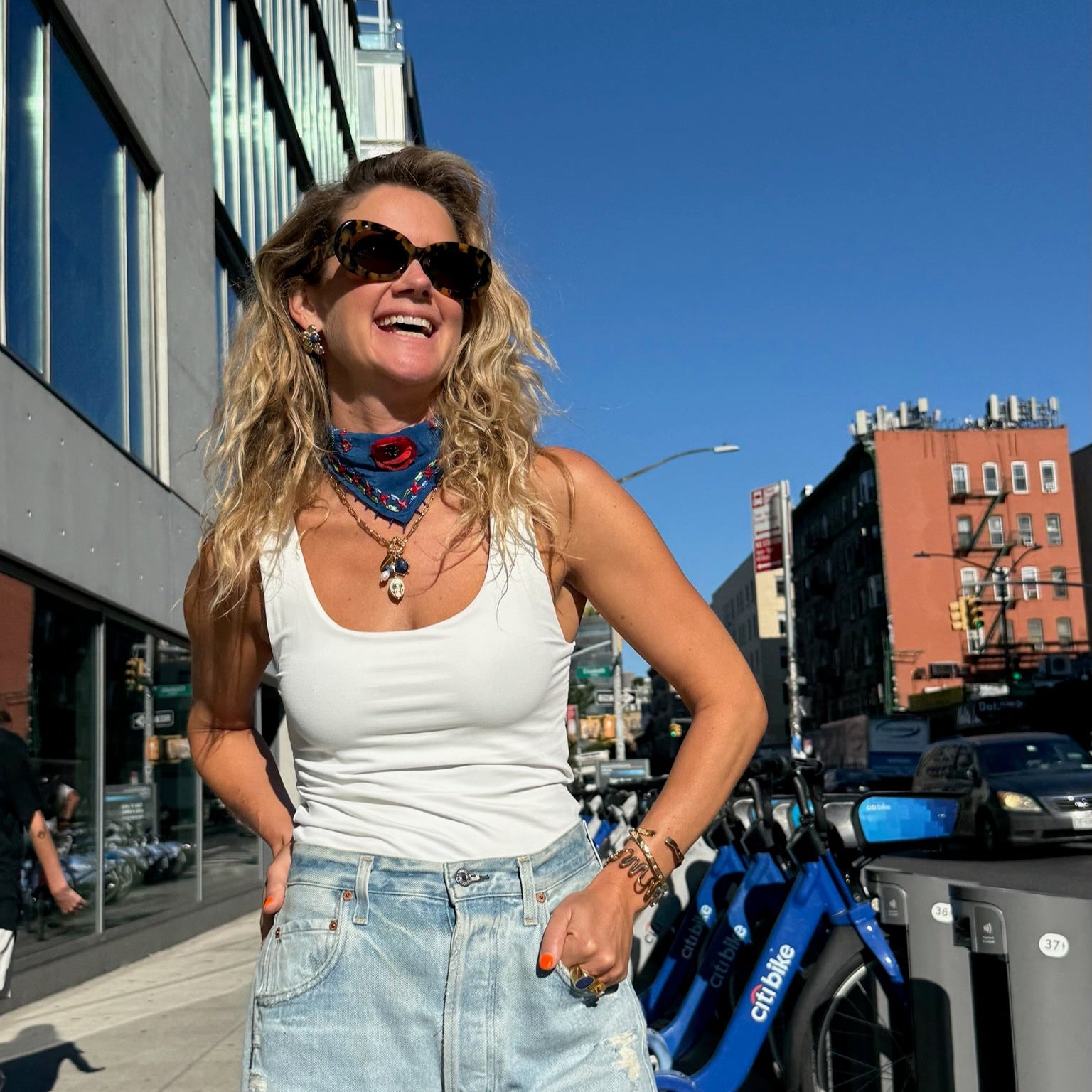 Maggie standing on a city street with buildings and clear blue sky in the background wearing blue bandana and charm necklace