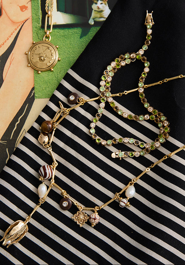 green and brown tennis necklace laying on top of a gold charm necklace against a striped background