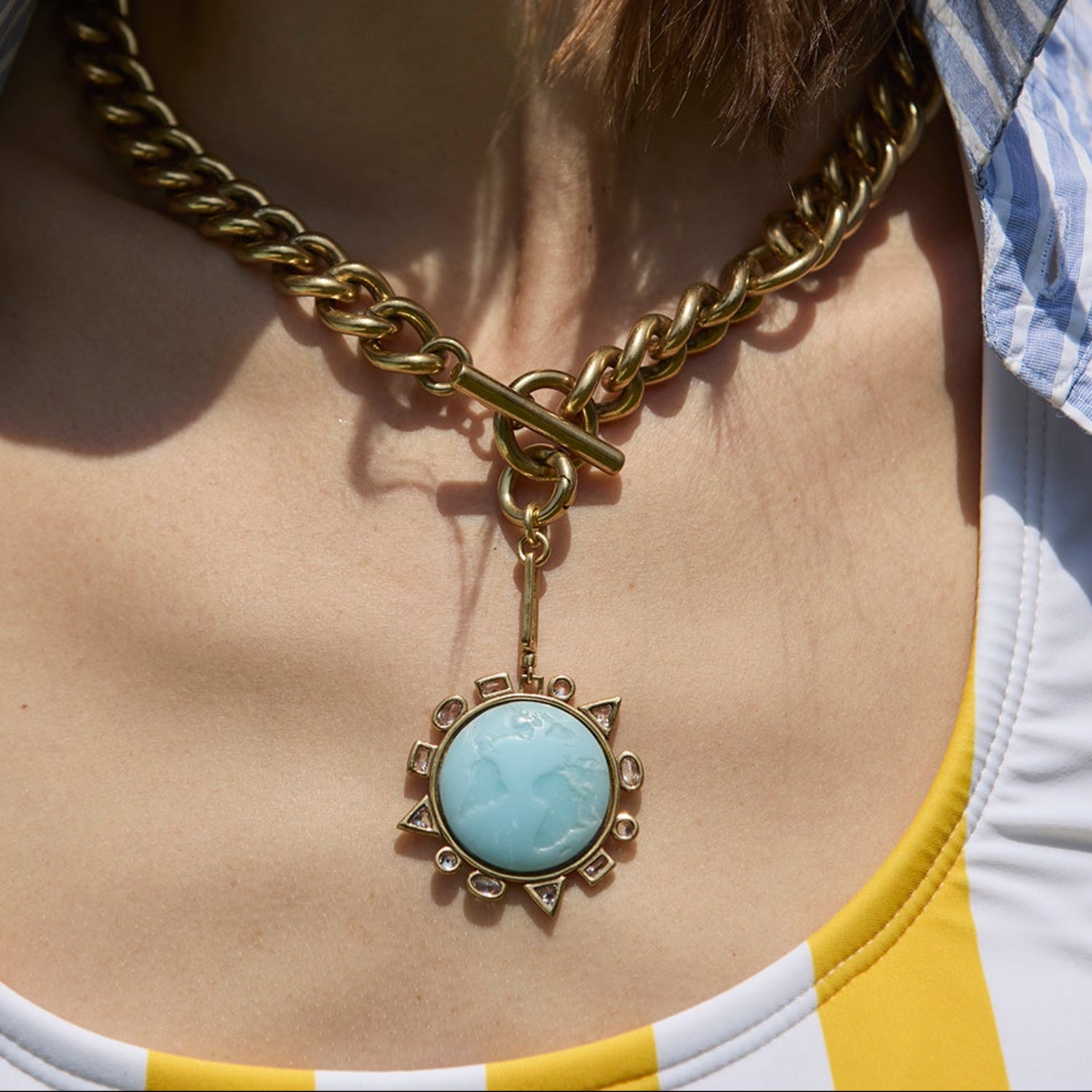 Person wearing a gold chain necklace with a blue pendant, wearing a striped shirt and yellow top.
