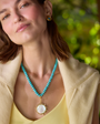 A woman wearing a turquoise beaded necklace with a large oval white pendant framed in gold, styled with a yellow top and beige jacket against a blurred green background.