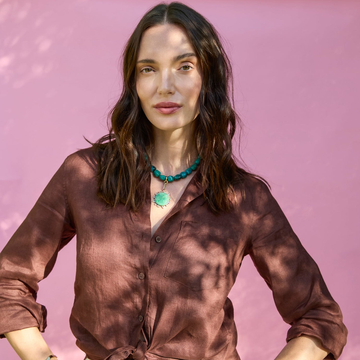 Woman wearing a brown blouse, chunky green necklace with charm against a green background