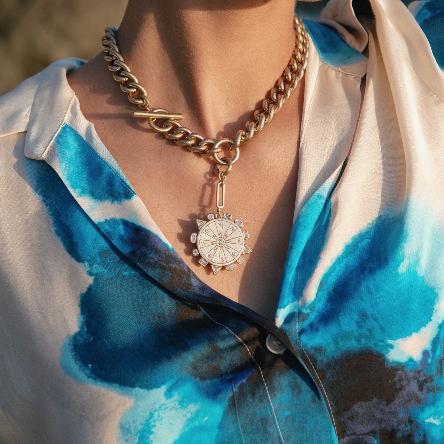 Person wearing a gold chain necklace with a pendant, set against a blurred natural background.