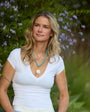 A woman with blonde hair wearing a white V-neck t-shirt and a turquoise beaded necklace with a white pendant, standing outdoors in front of green foliage and purple flowers.