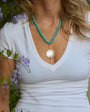A woman wearing a turquoise beaded necklace with a round white pearl pendant set in a rose gold floral frame, styled with a white V-neck t-shirt outdoors among green leaves and purple flowers.
