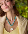 A woman wearing a turquoise beaded necklace with a large white shell pendant in a gold frame, styled with a yellow top and beige cardigan against a blurred green background.