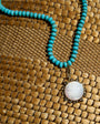 A turquoise beaded necklace with a round white mother-of-pearl pendant set in an ornate bronze-toned frame, displayed on a woven brown mat.