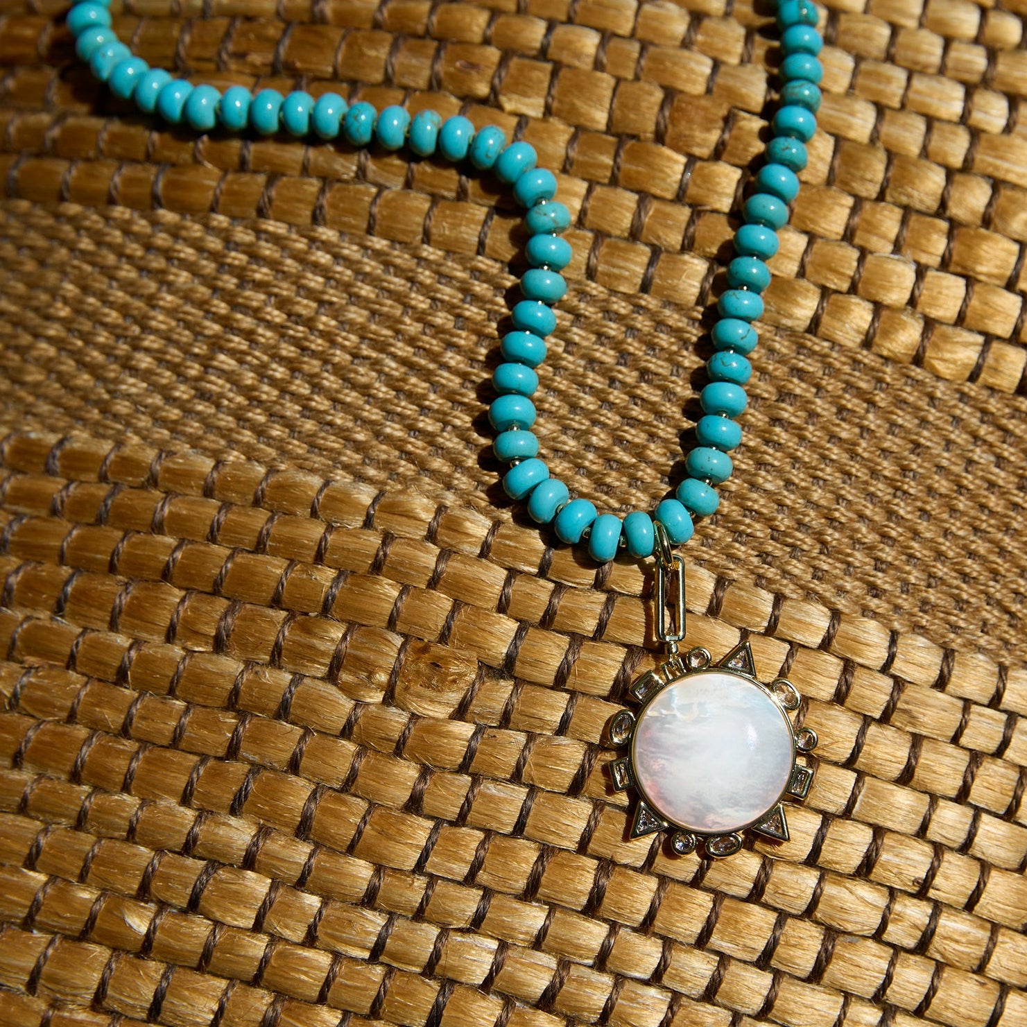 A turquoise beaded necklace with a round white mother-of-pearl pendant set in an ornate bronze-toned frame, displayed on a woven brown mat.