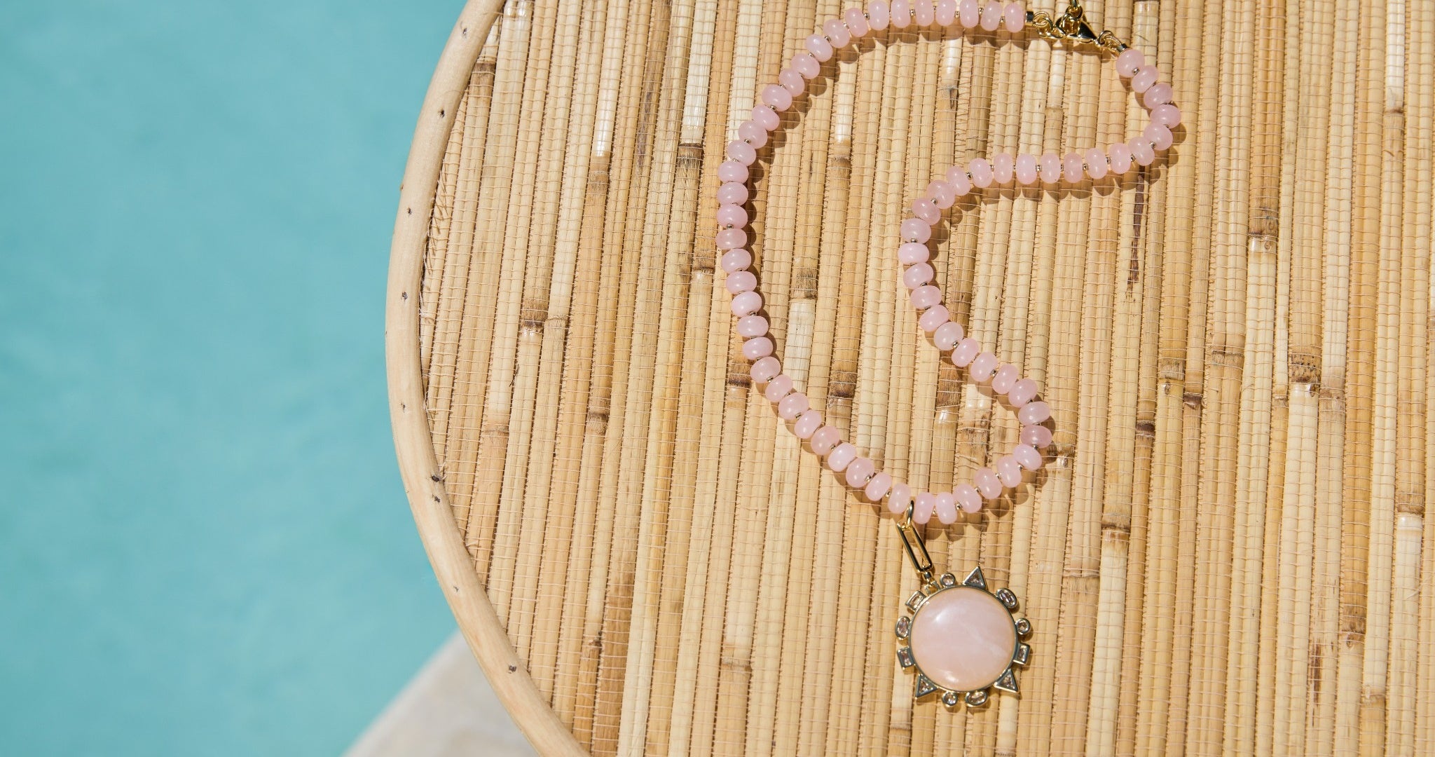 A pink beaded necklace with a round pink stone pendant in a gold-toned setting, arranged on a round woven mat next to a turquoise blue surface.