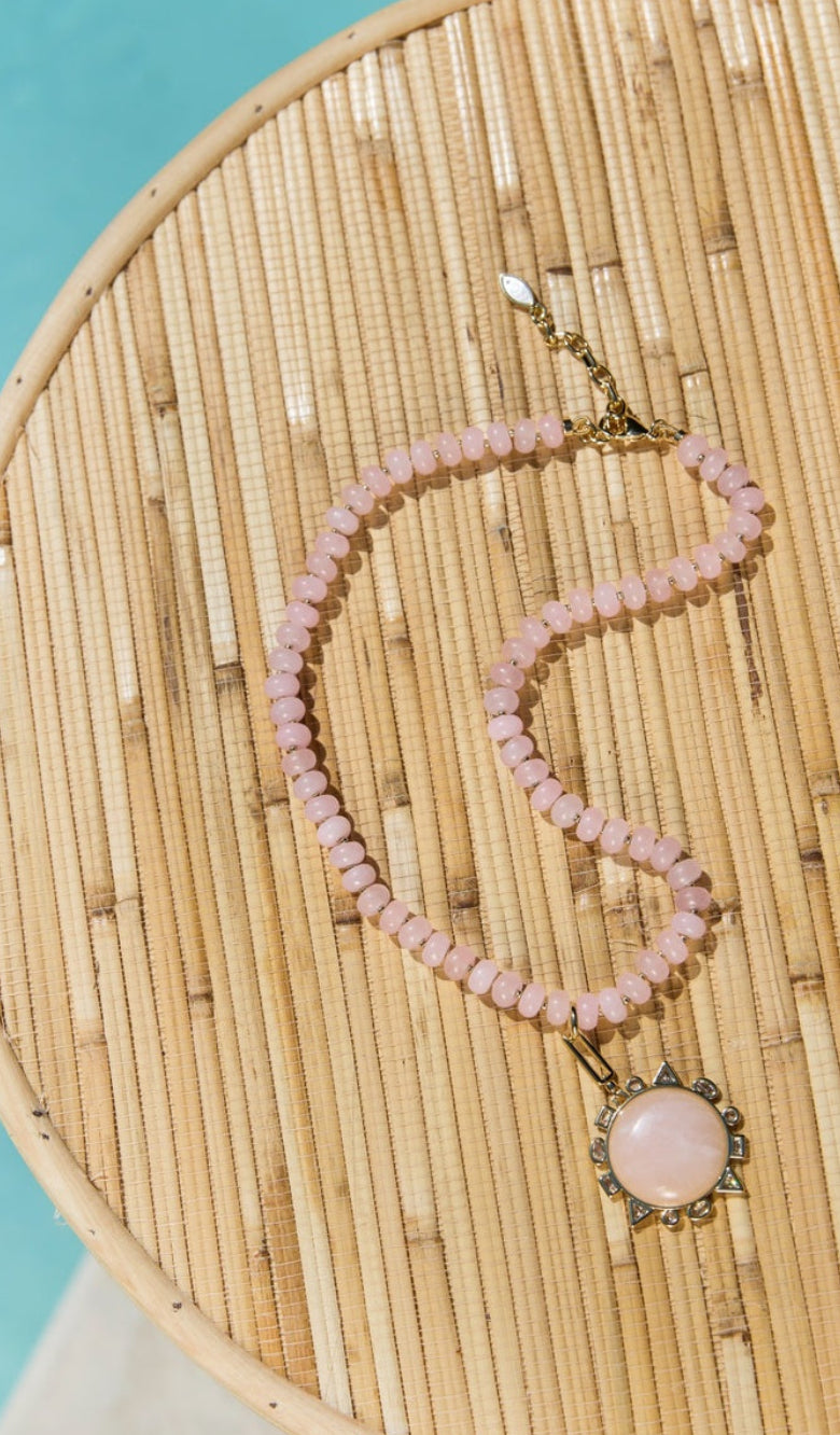 A pink beaded necklace with a rose quartz pendant and gold-tone clasp, displayed on a round woven bamboo tray against a turquoise background.