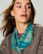 A woman wearing a teal and green patterned scarf with gold drop earrings, styled with a brown knit sweater against a light gray background.