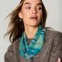 A woman wearing a teal and green patterned scarf with gold drop earrings, styled with a brown knit sweater against a light gray background.