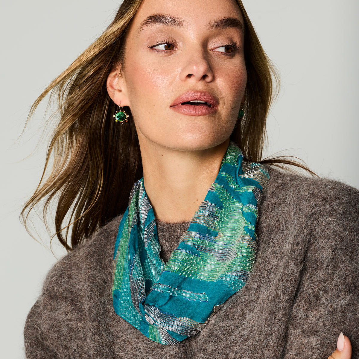 A woman wearing a teal and green patterned scarf with gold drop earrings, styled with a brown knit sweater against a light gray background.
