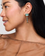 Woman wearing a fish-shaped earring with a green gemstone, Paula Earrings, on a plain background