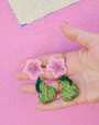 Hand holding green embroidered earrings with pink floral elements against a pink background