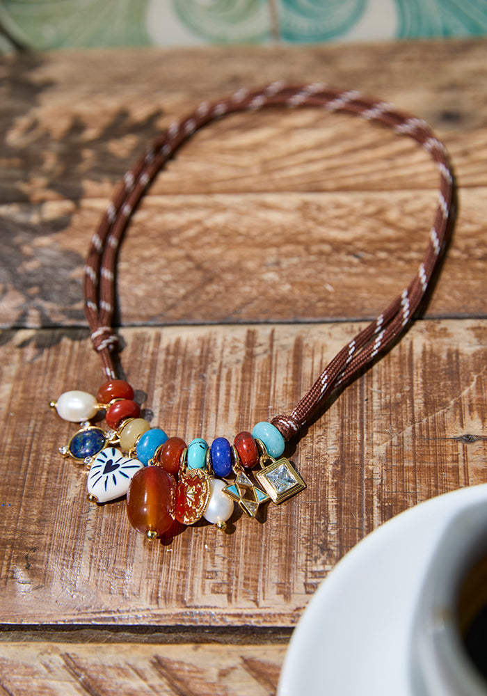 A bohemian-style beaded necklace with a brown braided cord and a cluster of colorful glass beads, charms, and stones, displayed on a rustic wooden table.