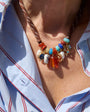 A close-up of a bohemian-style beaded necklace worn by a person in a white button-down shirt with thin red stripes, under bright natural sunlight.