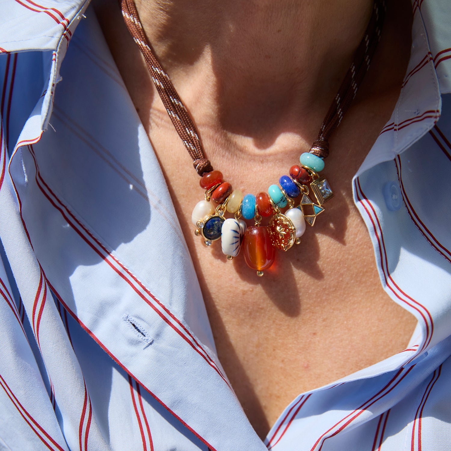 A close-up of a bohemian-style beaded necklace worn by a person in a white button-down shirt with thin red stripes, under bright natural sunlight.