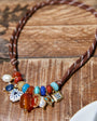 A bohemian-style beaded necklace with a brown braided cord and a cluster of colorful stones, pearls, and charms, displayed on a rustic wooden surface.