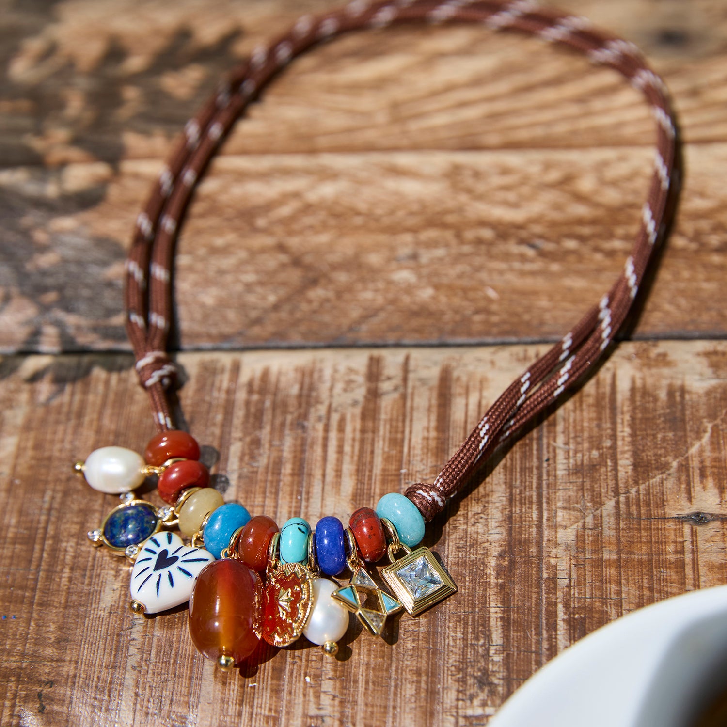 A bohemian-style beaded necklace with a brown braided cord and a cluster of colorful stones, pearls, and charms, displayed on a rustic wooden surface.