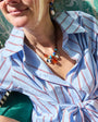 A close-up lifestyle shot of a woman wearing a light blue and white striped shirt with thin red stripes, accessorized with a bohemian beaded necklace and earrings, sitting in a green patterned chair.