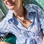 A close-up lifestyle shot of a woman wearing a light blue and white striped shirt with thin red stripes, accessorized with a bohemian beaded necklace and earrings, sitting in a green patterned chair.