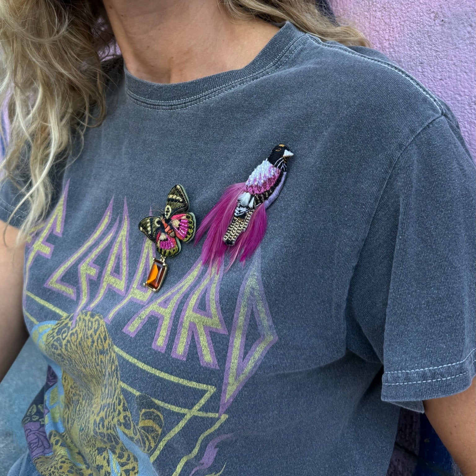 Person wearing a graphic t-shirt with colorful designs  wearing embroidered brooches against a graffiti wall.