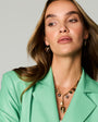 A close-up of a woman wearing gold heart-shaped dangle earrings and a gold pendant necklace, styled with a mint green blazer against a light gray studio background.