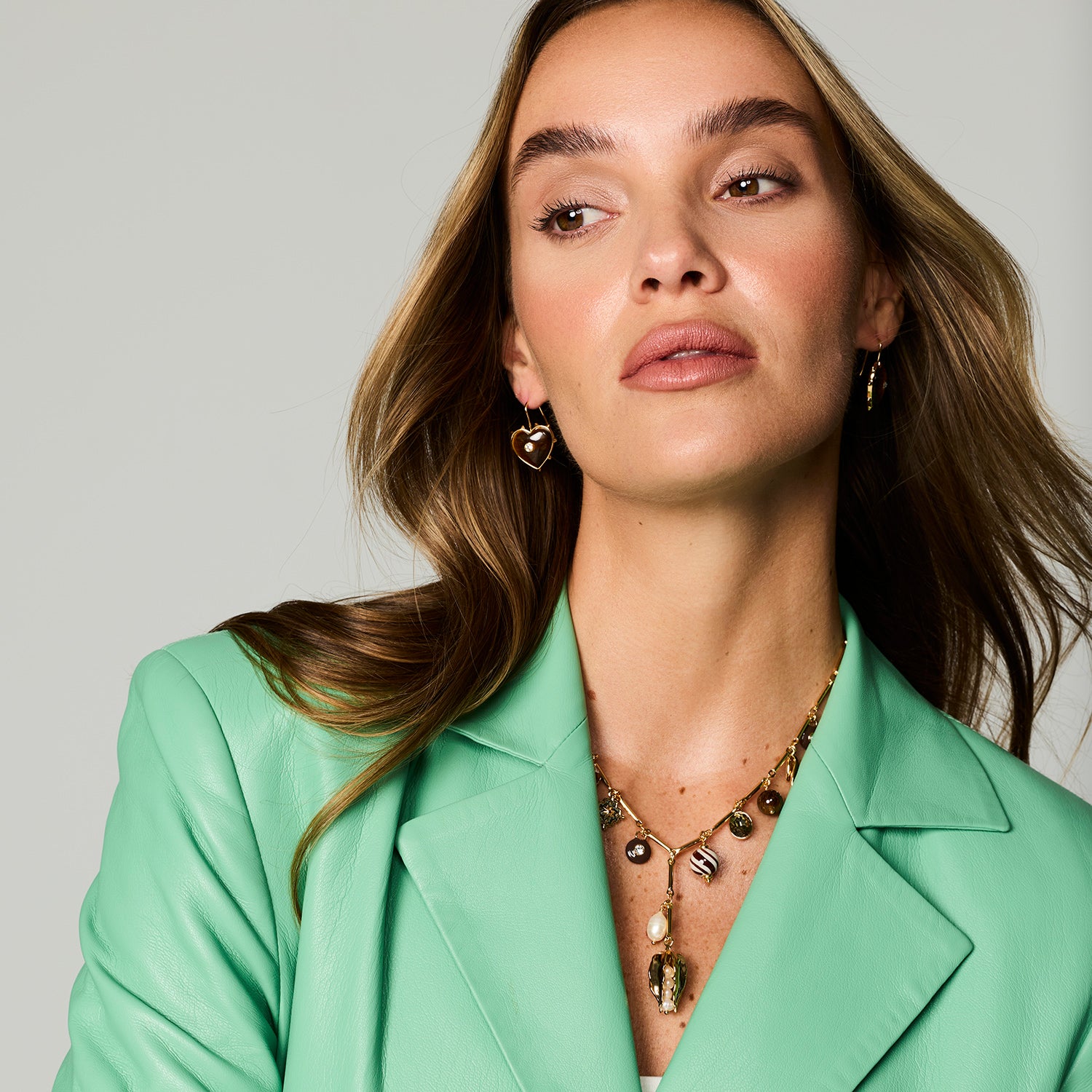 A close-up of a woman wearing gold heart-shaped dangle earrings and a gold pendant necklace, styled with a mint green blazer against a light gray studio background.
