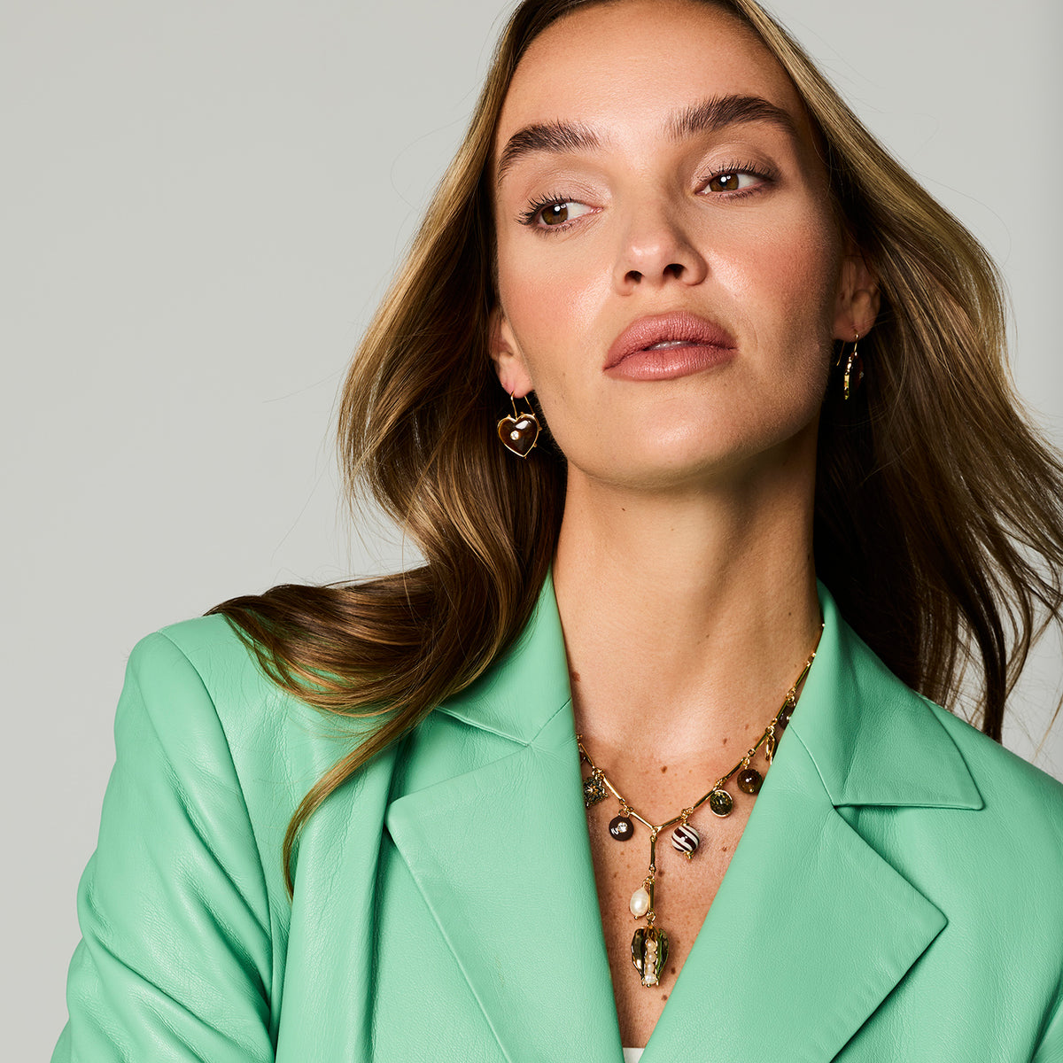 A close-up of a woman wearing gold heart-shaped dangle earrings and a gold pendant necklace, styled with a mint green blazer against a light gray studio background.