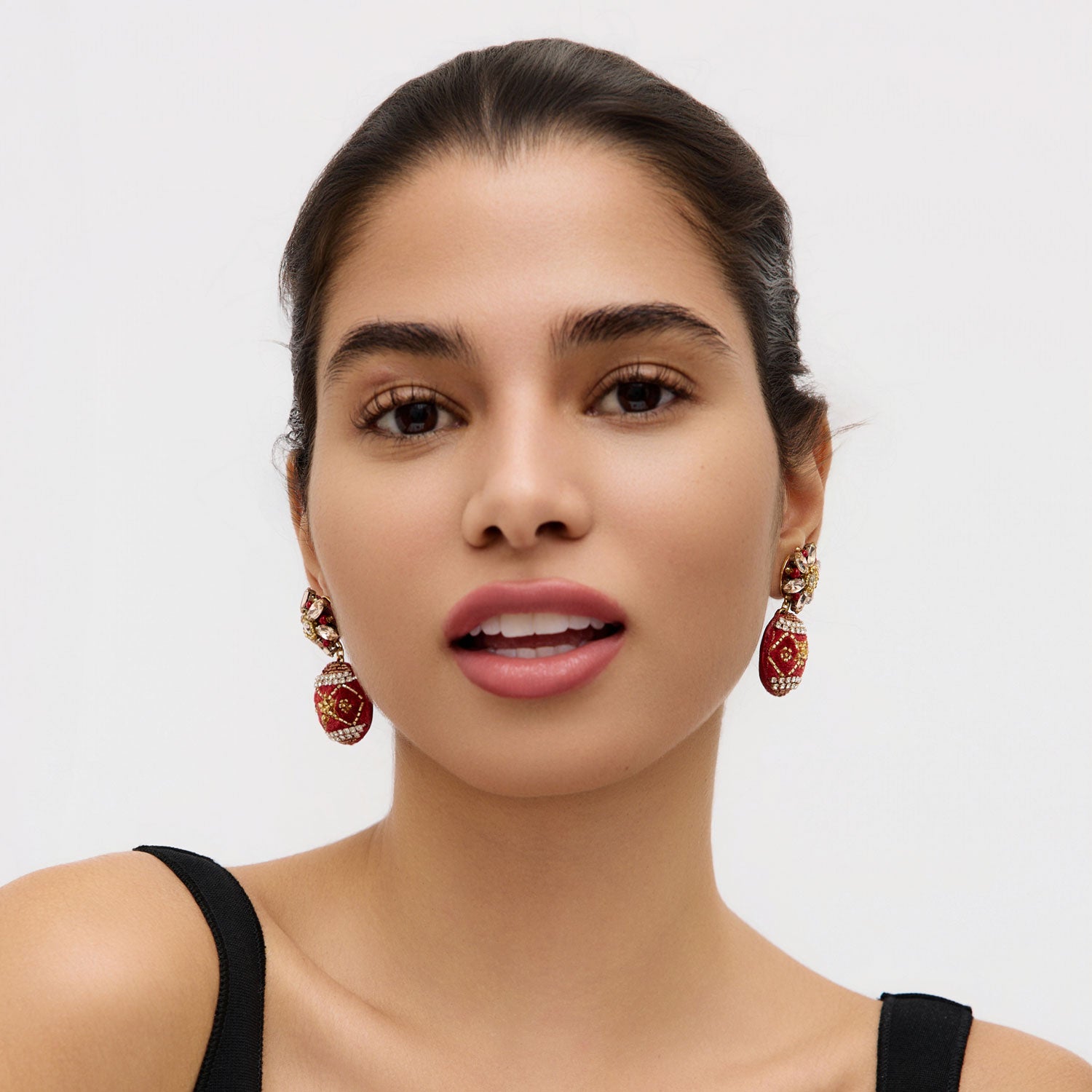 Woman wearing red ornament drop earrings against a plain background