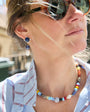 A close-up lifestyle shot of a woman wearing a colorful beaded necklace and matching drop earrings with blue and white stones.