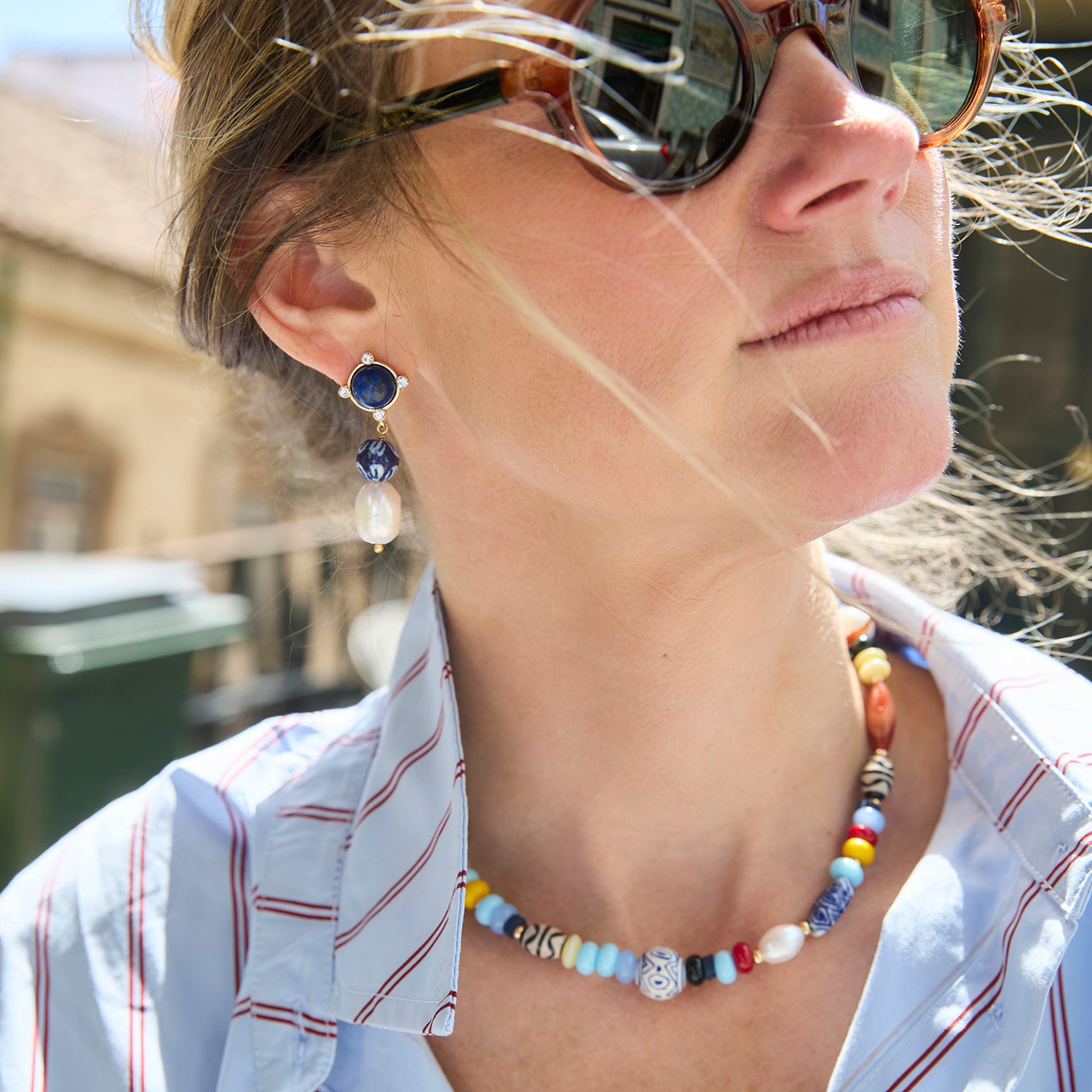 A close-up lifestyle shot of a woman wearing a colorful beaded necklace and matching drop earrings with blue and white stones.