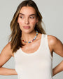 A woman wearing a colorful beaded necklace and a white tank top, photographed against a plain light gray studio background.