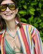 Woman wearing sunglasses, a gold charm necklace and turquoise beaded necklace with a colorful striped shirt with a green leafy background