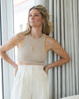 Woman wearing a beige top and white pants with a gold chain and charm necklace around neck standing indoors with curtains in the background