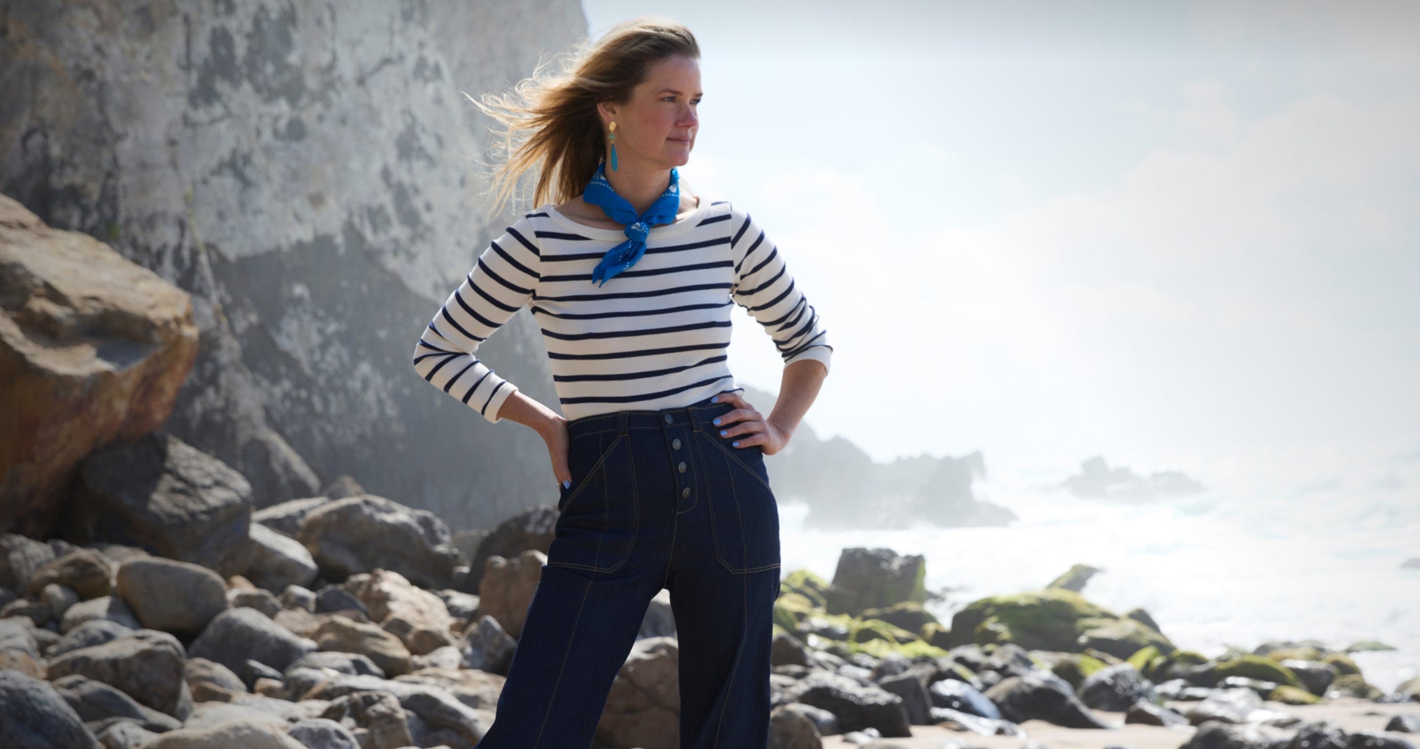 A woman stands on a rocky beach wearing a navy and white striped long-sleeve top with a bright blue neck scarf and dark wide-leg jeans, with a large rock formation and sea in the background.
