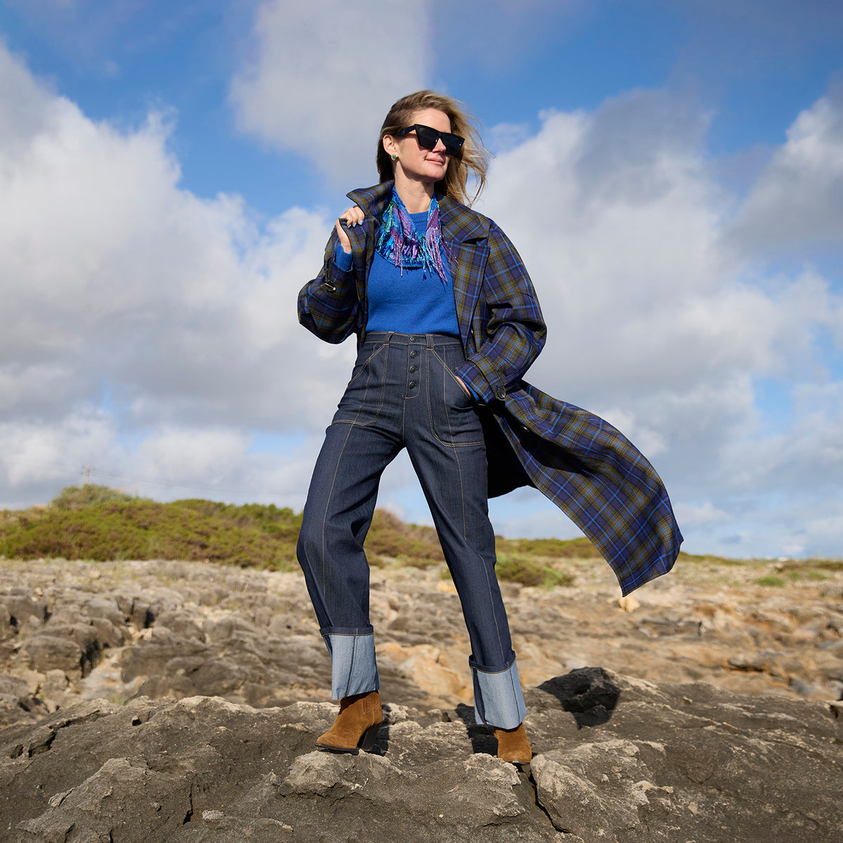 Model wearing a long plaid jacket and denim jeans with a colorful scarf necklace around neck against a rocky background