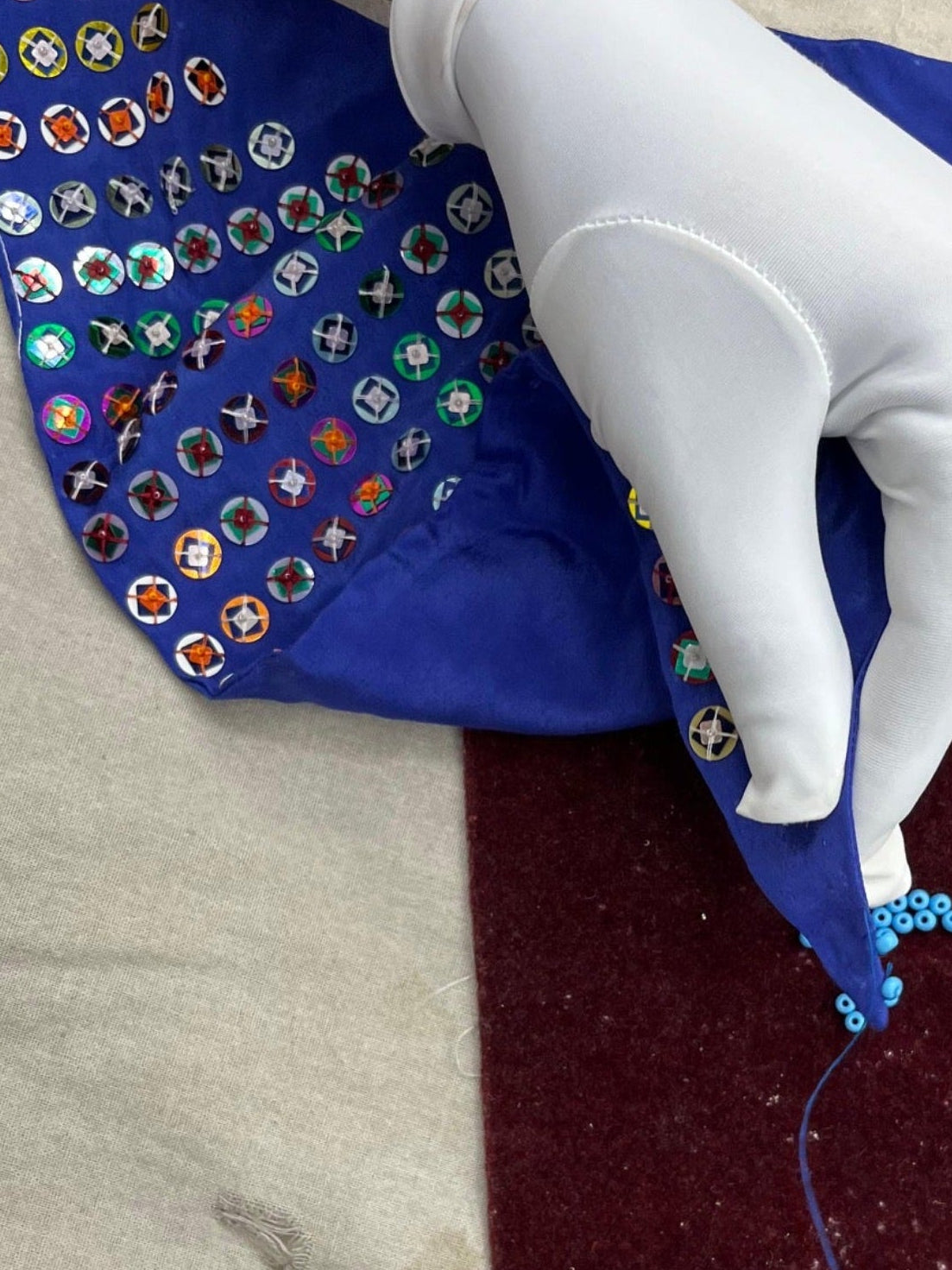 A close-up of a hand wearing a white glove holding a blue fabric piece decorated with many colorful embroidered or applique motifs, while placing it over a dark burgundy fabric surface on a light beige cloth background.