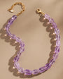 A beaded necklace made of oval translucent purple gemstone beads with a gold-tone chain and lobster clasp, arranged on a warm beige background with soft shadows and light flares.