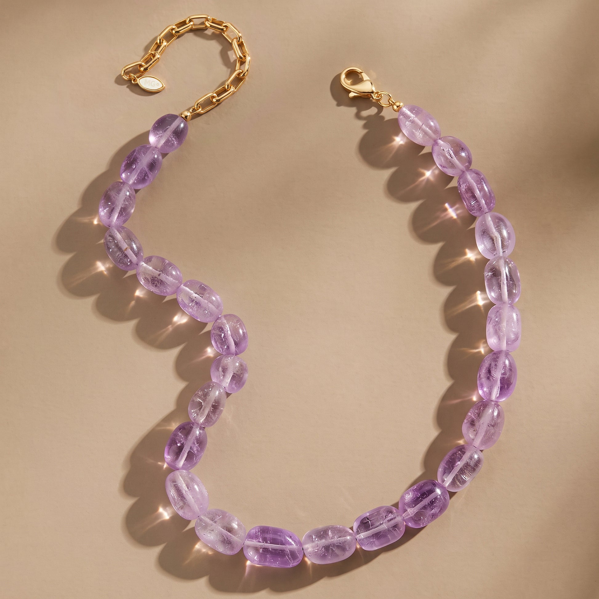 A beaded necklace made of oval translucent purple gemstone beads with a gold-tone chain and lobster clasp, arranged on a warm beige background with soft shadows and light flares.