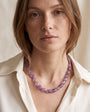 A close-up of a woman wearing a chunky, translucent purple gemstone beaded necklace with a cream-colored collared shirt.