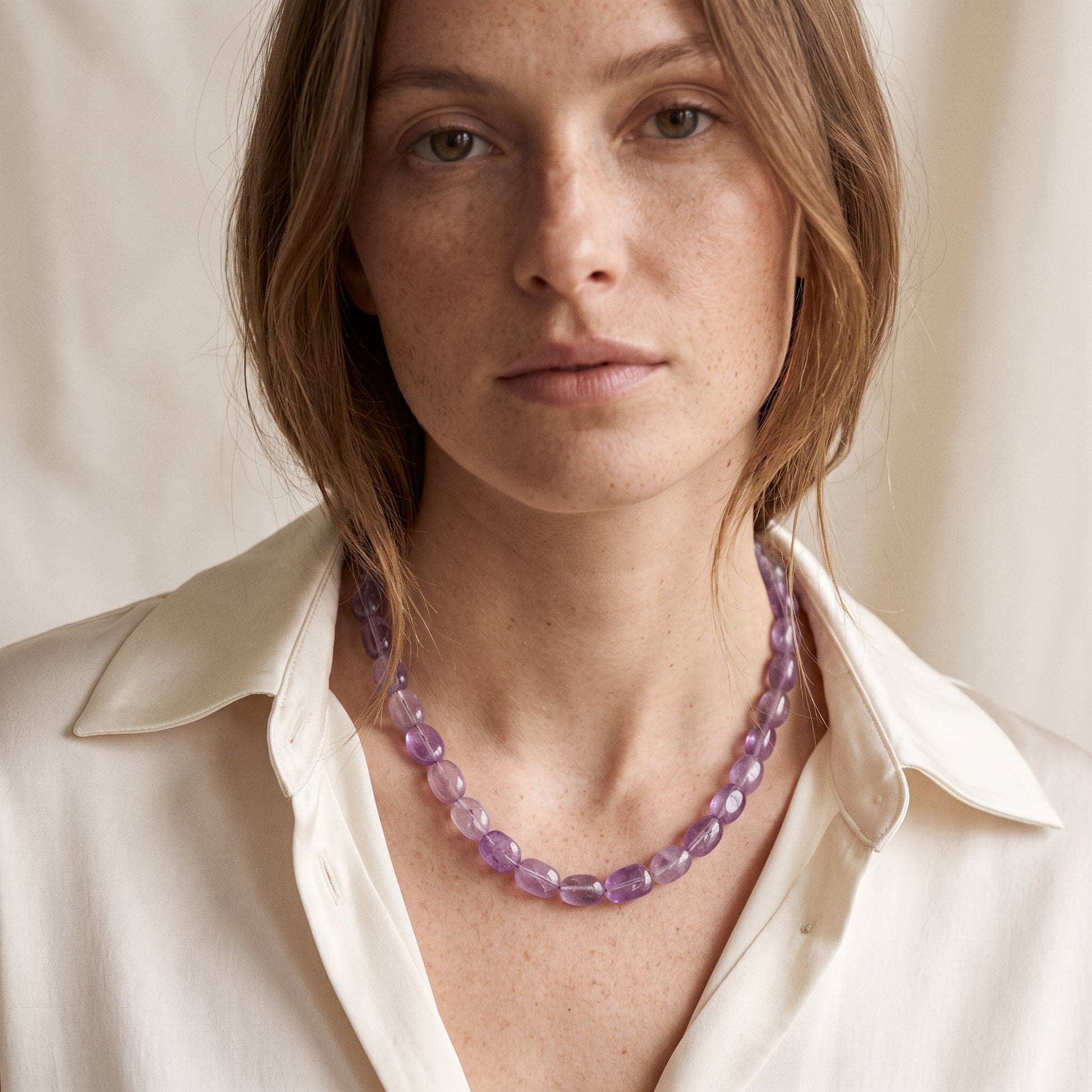 A close-up of a woman wearing a chunky, translucent purple gemstone beaded necklace with a cream-colored collared shirt.