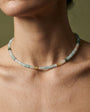 A close-up of a person's neck and upper chest wearing a delicate beaded choker necklace. The necklace features small translucent beads in pale blue, white, and a few warm gold and green accents, set against a soft olive green background.